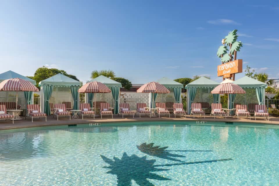 Resort pool with palm shadow reflection, red striped umbrellas, mint cabanas, and vintage signage