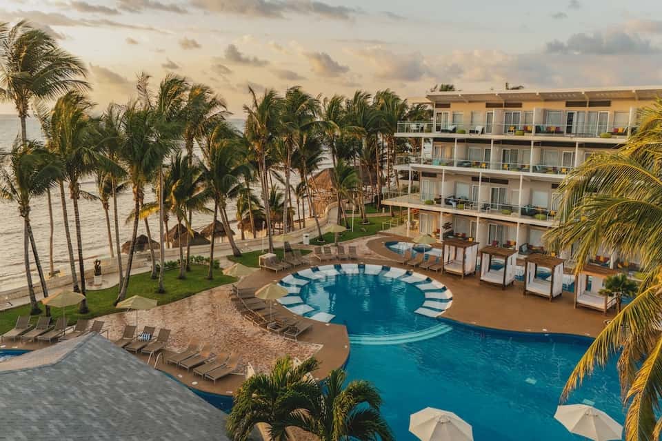 Beachfront resort with curved pool, palm trees, multi-story buildings, and ocean view at sunset