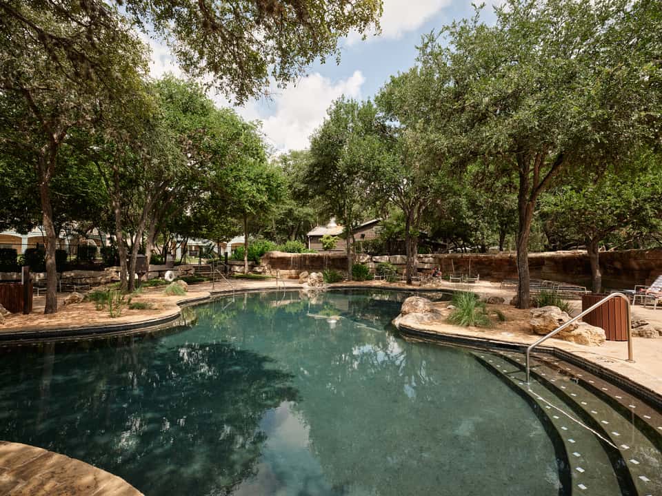 Natural spring-fed swimming pool surrounded by mature trees, stone features, and rustic stone buildings