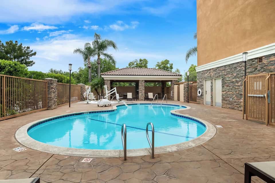 Resort swimming pool with lounge chairs, covered pavilion, stone patio, and palm trees under blue sky