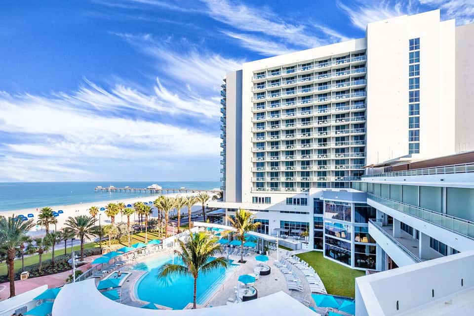 Beachfront hotel with white high-rise tower, curved blue pool, palm trees, and ocean view with pier