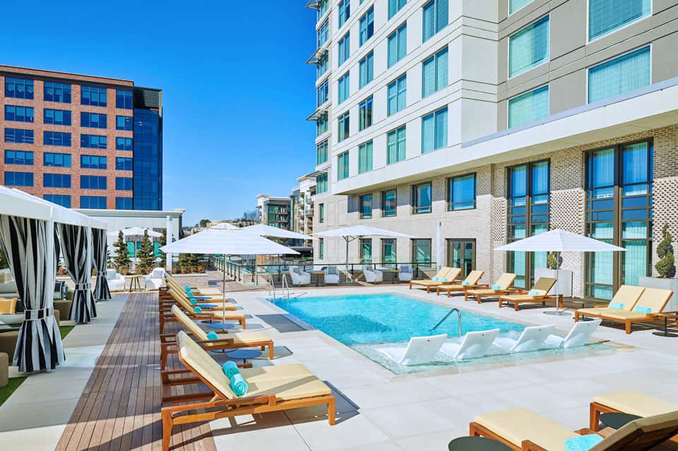 Modern resort pool deck with turquoise water, wooden loungers, white umbrellas, and multi-story buildings