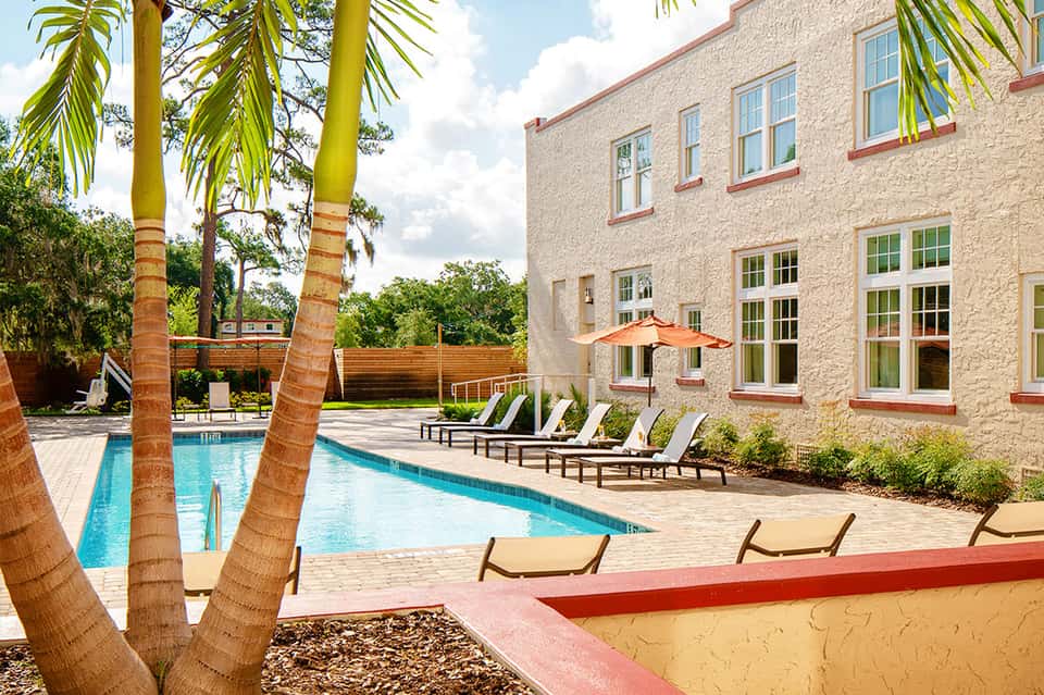 Resort exterior with swimming pool, palm trees, stone building with large windows, and lounge chairs under umbrellas