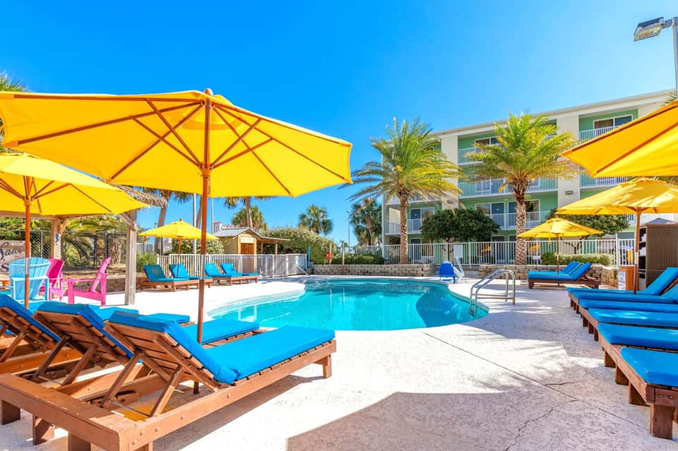 Resort pool area with bright yellow umbrellas, blue loungers, and palm trees under clear sky