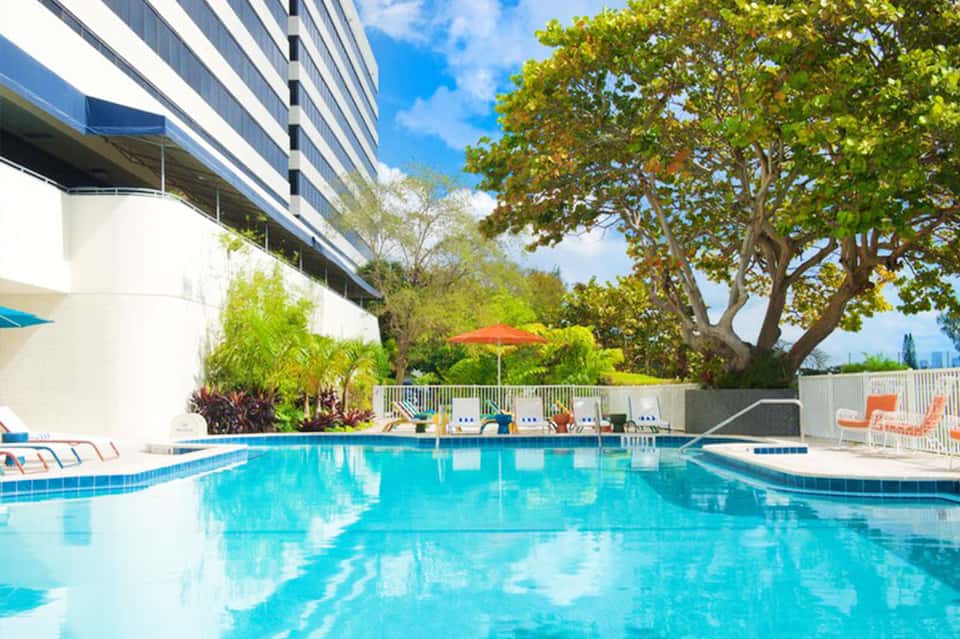 Resort swimming pool with lounge chairs, umbrellas, tropical landscaping, and multi-story building