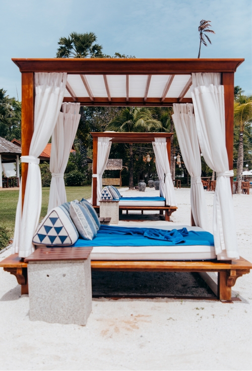A tranquil bed with blue and white curtains, perfectly placed on the sandy beach.