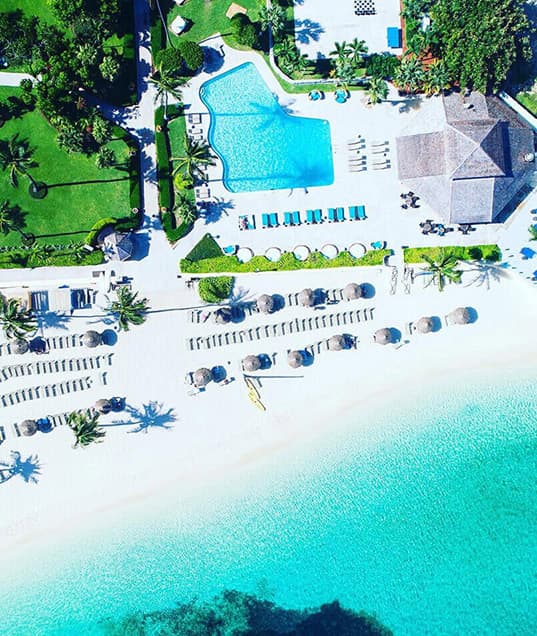 Aerial view of a hotel's private beach lined with lounge chairs and cabanas facing a clear aqua ocean