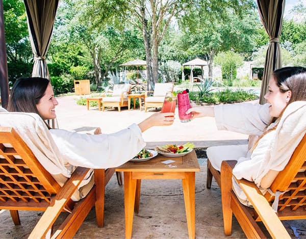 Two women doing a cheers while eating lunch under a cabana at a resort covered with trees