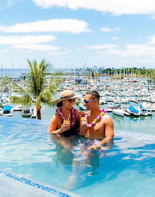 A couple drinking cocktails in a resort's infinity pool with a view of a harbor